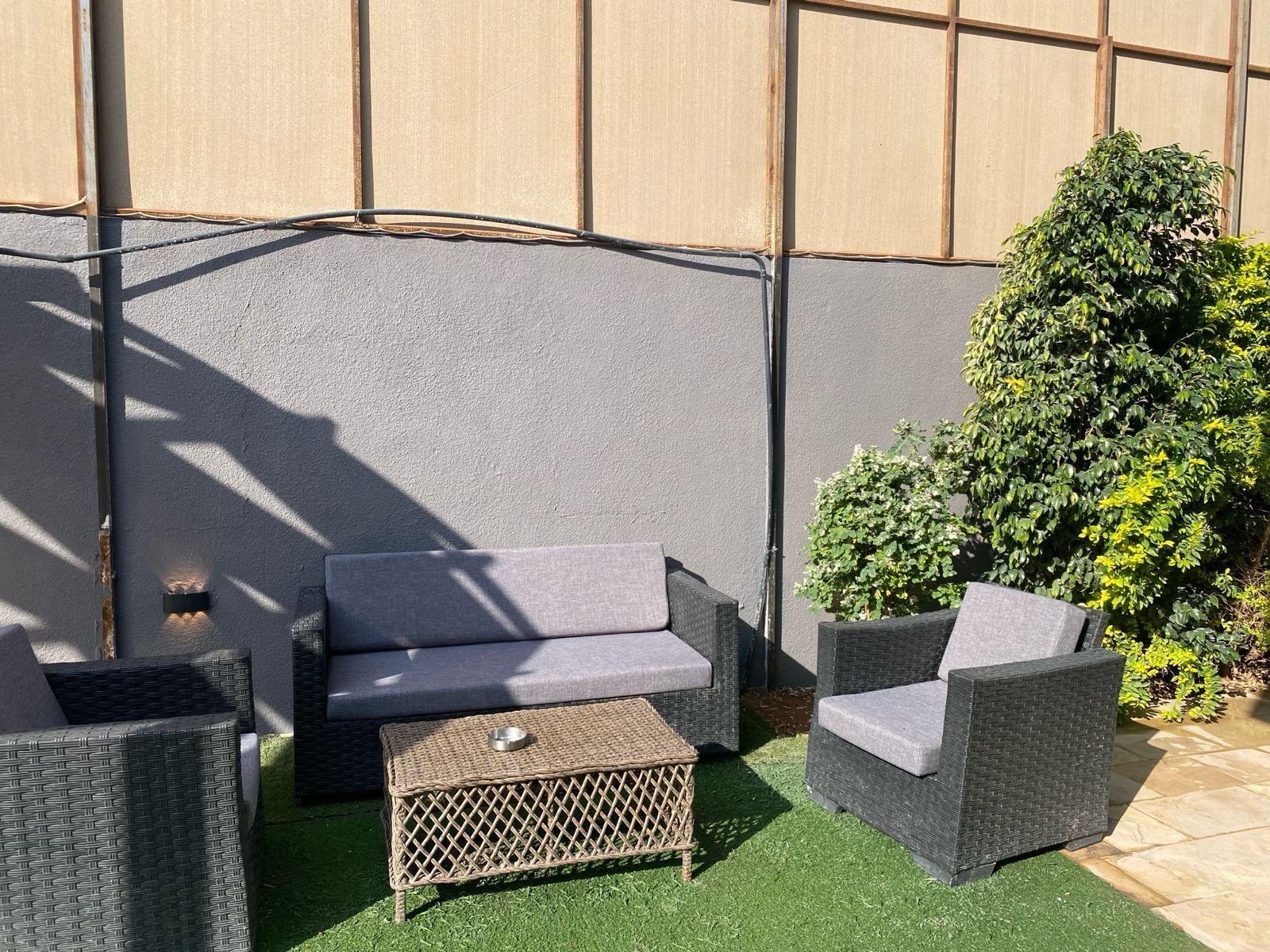 An outdoor seating area with wicker furniture and a small table, surrounded by green artificial grass and a backdrop of a grey wall with windows.