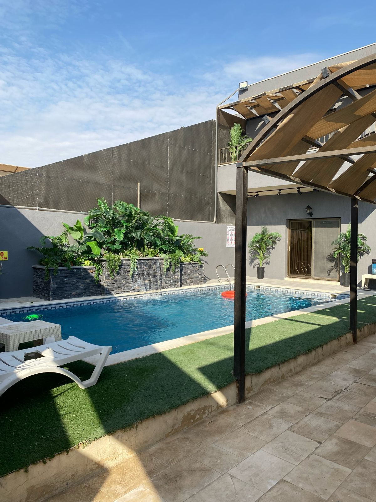 A swimming pool with a pergola, artificial grass, and a stone wall with plants.