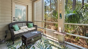 A covered porch with a wicker sofa and a small table, overlooking a forested area. - 55-2 Low Country Get-a-way 2 Bedroom Condo (Pawleys Island)