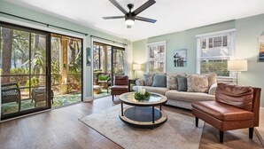 A spacious living room with a large sliding glass door leading to a garden, a ceiling fan, a round coffee table, and a sofa with multiple cushions. - 55-2 Low Country Get-a-way 2 Bedroom Condo (Pawleys Island)