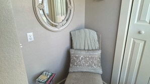 Una habitación pequeña y acogedora con un espejo redondo, un banco con cojín, una mesita con libros y una puerta.