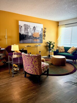 Un salon avec un tableau représentant un paysage urbain, une table basse ronde, un fauteuil violet, un canapé vert et une table basse en verre avec une lampe.