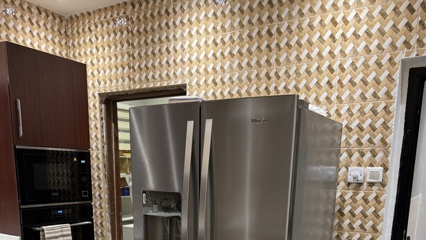 Un refrigerador de acero inoxidable con dispensador de agua, un microondas y un horno integrado en una cocina con paredes de baldosas estampadas y un gabinete de madera.