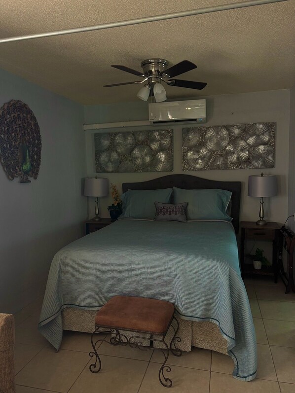 Un dormitorio con una cama, dos aires acondicionados instalados en la pared, un ventilador de techo, decoración en la pared y una mesita de noche con una lámpara.