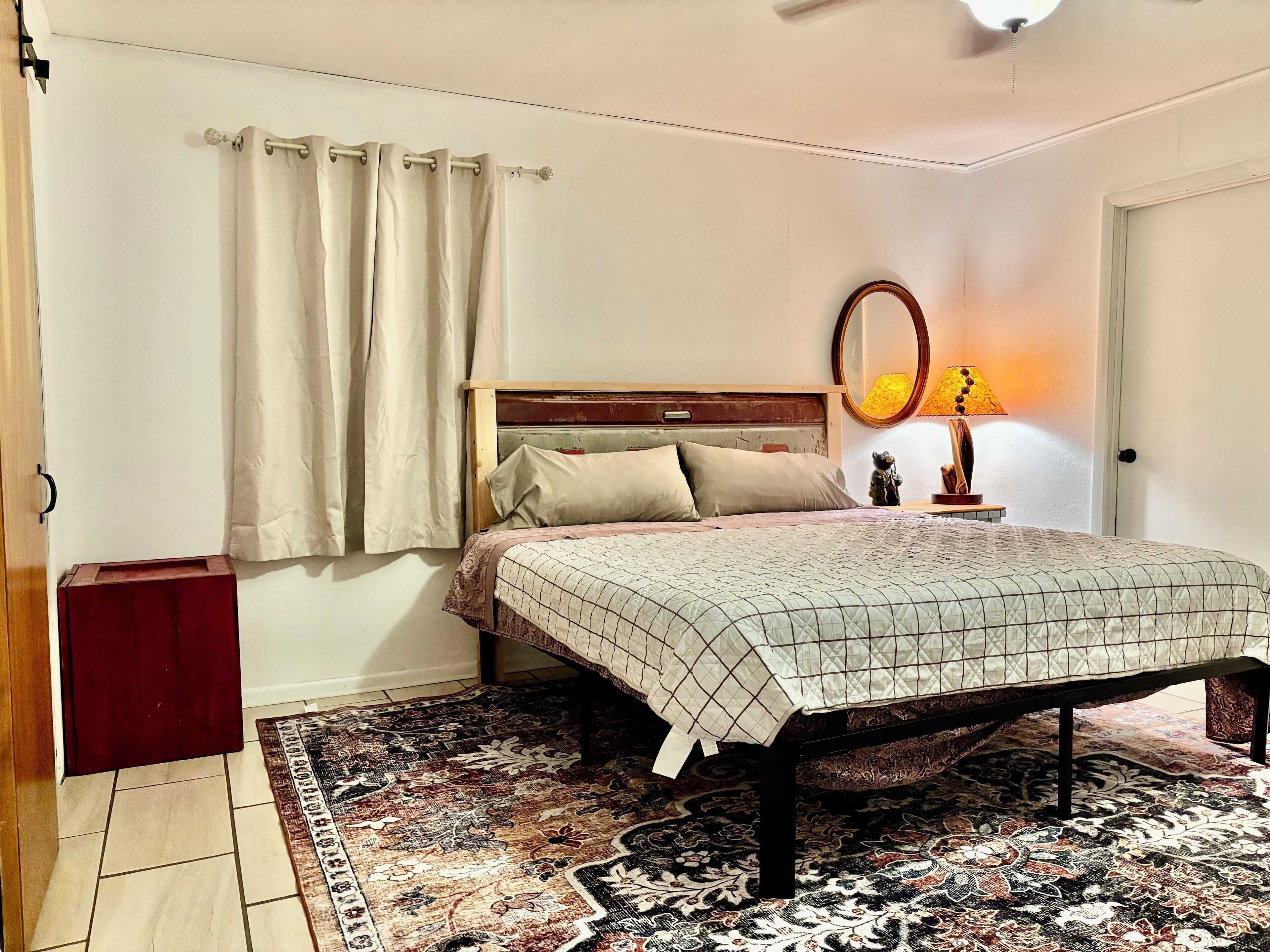 A bedroom with a bed, a nightstand, a lamp, a mirror, and a rug.