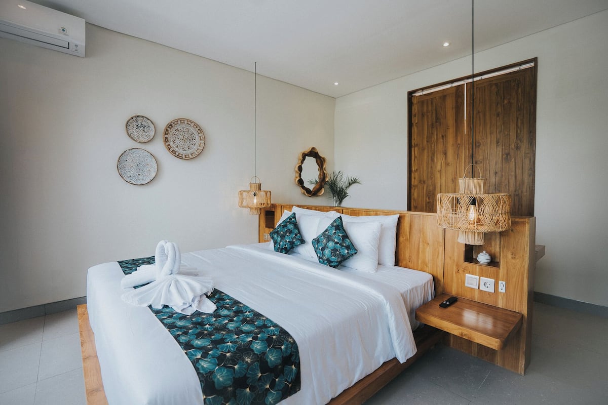 A neatly made bed with a white duvet and teal patterned throw, a wooden bedside table with a remote control, and a wooden wardrobe with a woven light fixture.