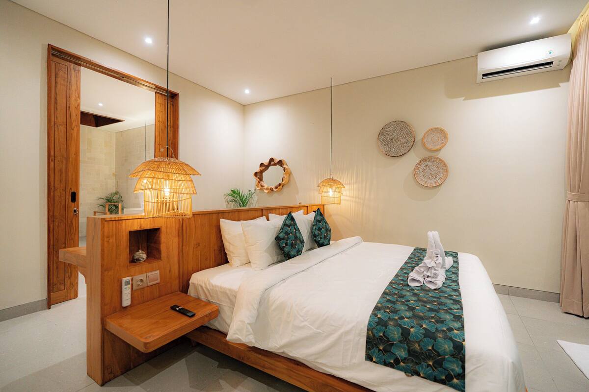A neatly arranged hotel room with a wooden headboard, a bedside table with a remote control, a bed with white bedding and a patterned throw, and wall-mounted decor including a mirror and wall art.