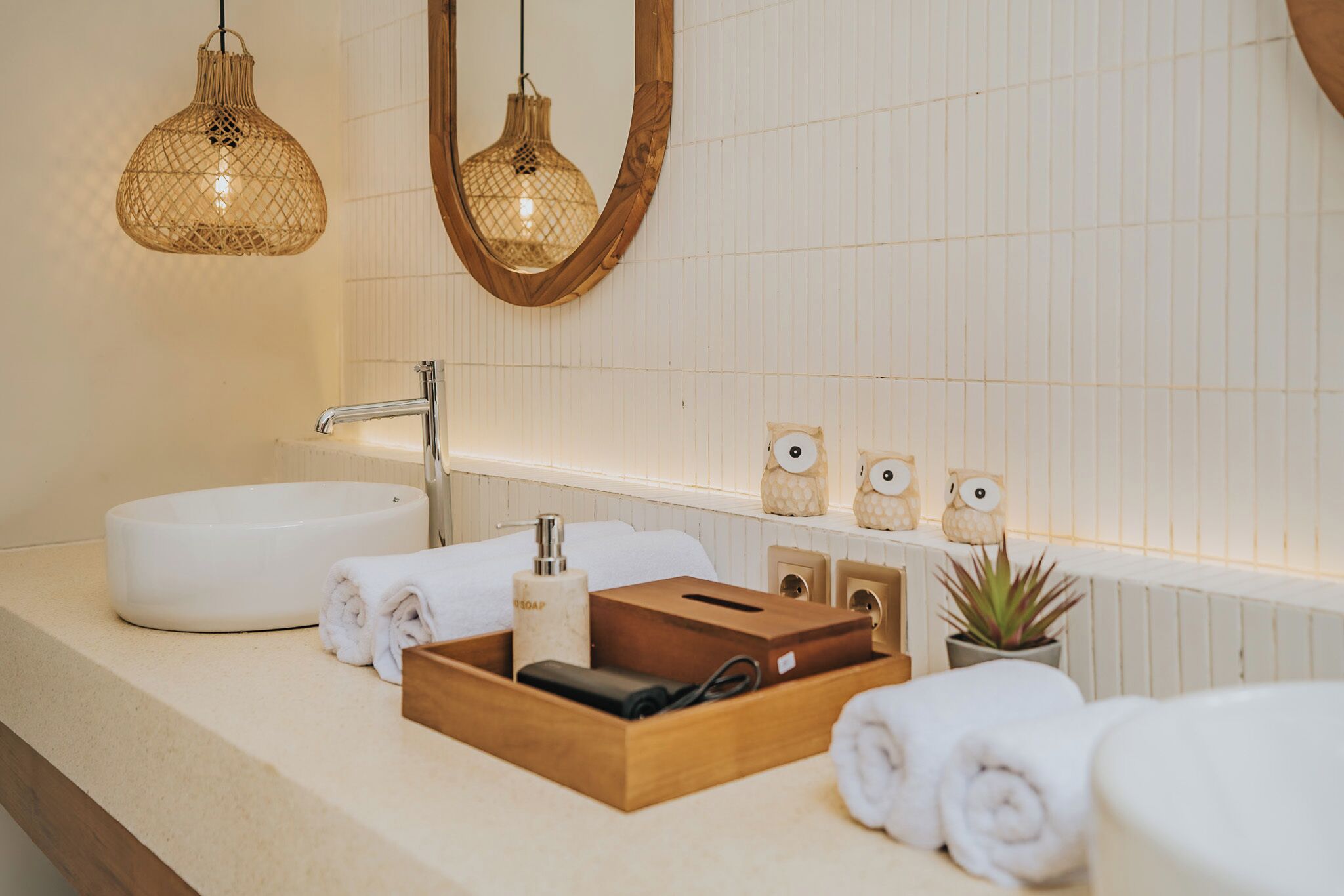 A bathroom with a white sink, wooden storage box, rolled towels, a soap dispenser, and two owl-shaped ornaments on a shelf.