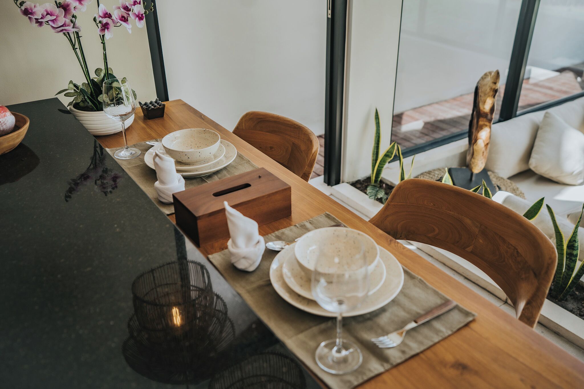 Een eettafel gedekt met borden, kommen en een wijnglas, omringd door stoelen en potplanten.