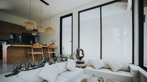 A modern living room with a white sofa, a wooden coffee table, and a kitchen area with wooden cabinets and a bar. - Villa Manna Ubud (Ubud)