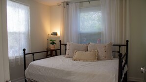 A neatly made bed with a white bedspread and several pillows, a bedside table with a lamp, and a window with blinds.
