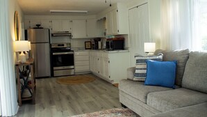 A compact kitchen with white cabinets, a refrigerator, stove, and microwave. There is a gray sofa with a blue pillow and a wooden side table with a lamp.