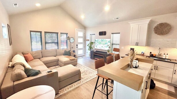 A spacious living area with a large L-shaped sofa, a dining table with bar stools, and a kitchen area with white cabinets and a sink.