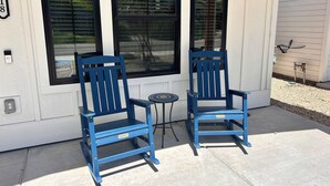 Two blue Adirondack chairs outside a house with large windows and a small table between them.