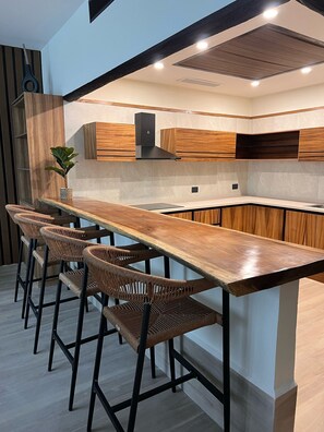 A modern kitchen with a long wooden bar, four wicker bar stools, and built-in wooden cabinets. - Hotel Mundo Maya Tulum Aeropuerto (Chunyaxché)