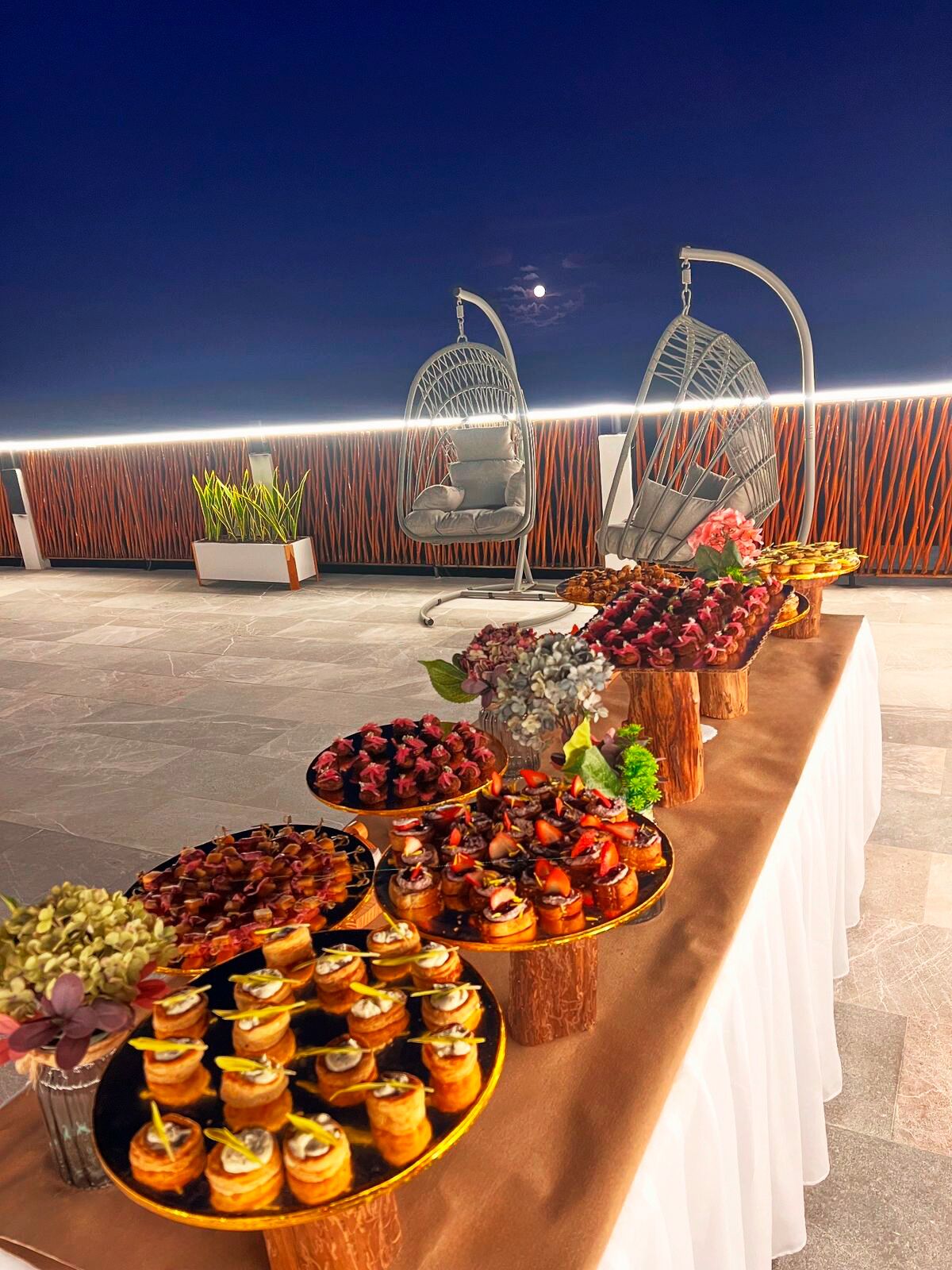 Un espacio de comedor en la azotea con diversos aperitivos y postres, dos sillones colgantes y vistas del cielo nocturno.