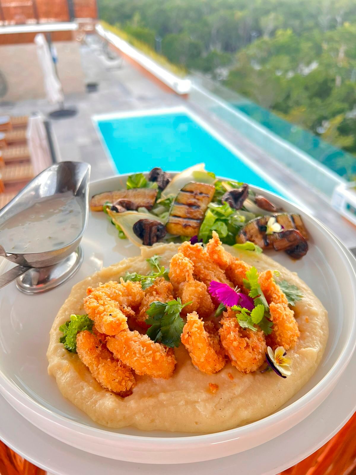A meal with breaded shrimp and grilled vegetables, served with a side of soup and salad, overlooking a pool and greenery.