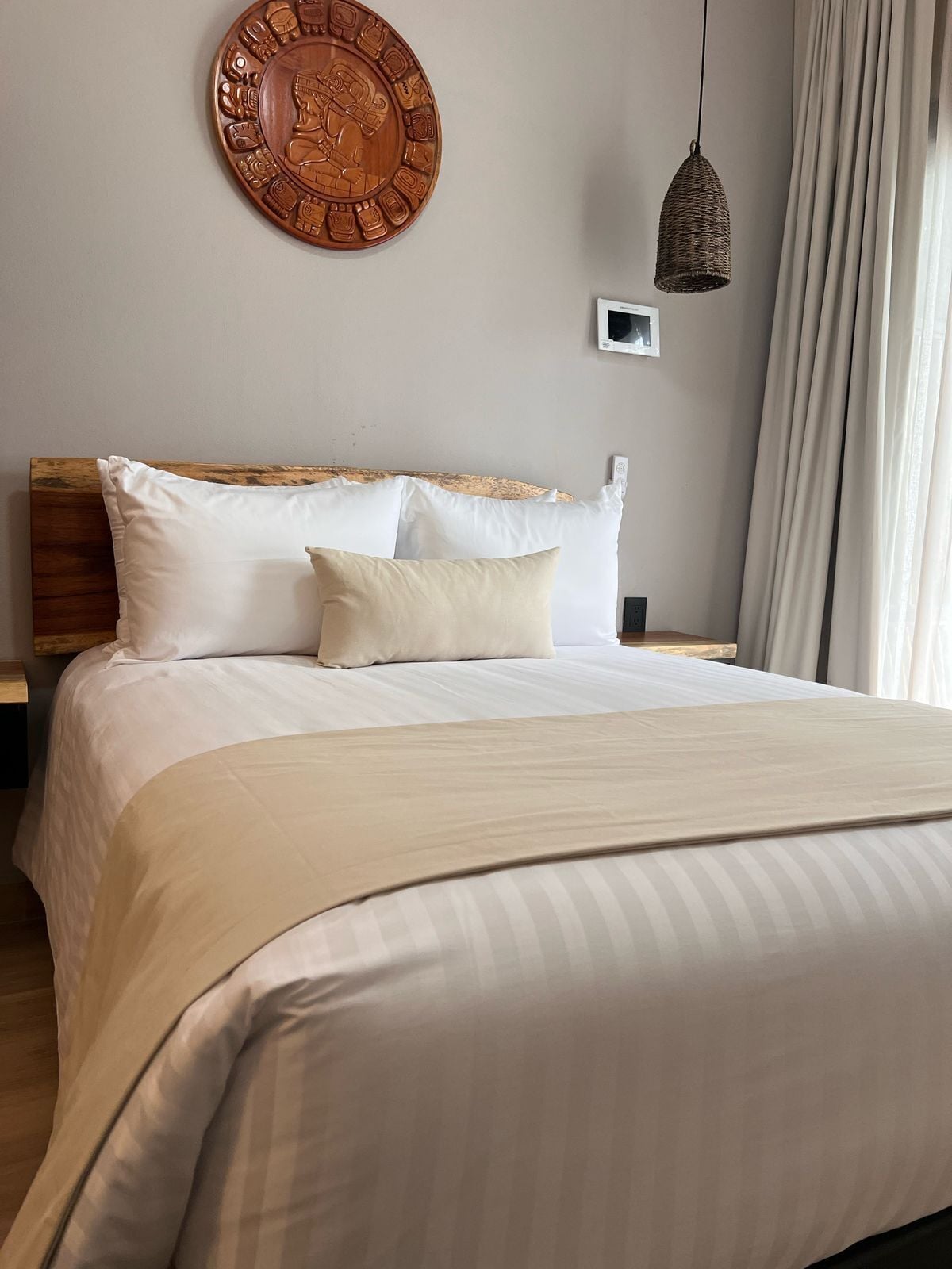 Una cama bien hecha con ropa de cama blanca y beige, un cabecero de madera y un adorno redondo montado en la pared.