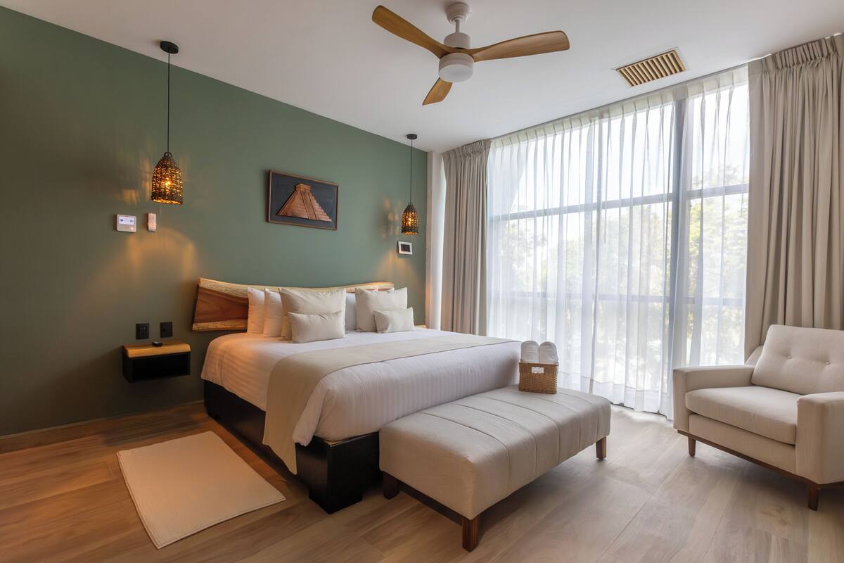 Ein Schlafzimmer mit einem Bett, einem Nachttisch, einer Bank, einem Sessel, einem Deckenventilator und großen Fenstern mit Vorhängen.