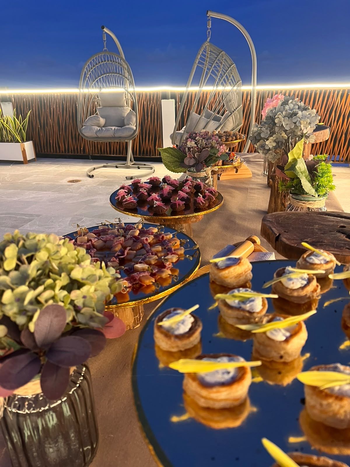 Un área de comedor al aire libre con diferentes alimentos en fuentes, rodeada de sillas y plantas en macetas.