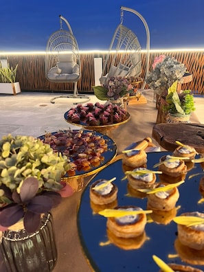 Un área de comedor al aire libre con diferentes alimentos en fuentes, rodeada de sillas y plantas en macetas.