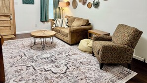 Un salón con alfombra estampada, un sofá, un sillón con estampado, una mesita redonda y una pared decorada con diversos objetos tejidos.
