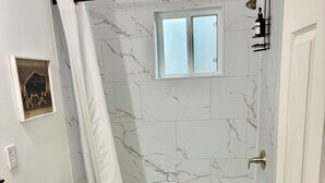 Un baño con ducha de azulejos de mármol, inodoro blanco, lavabo con tocador de madera y alfombra estampada en el suelo.