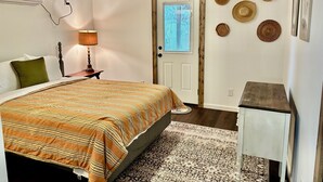Un dormitorio con cama, una mesita de noche con lámpara, ventilador de techo, puerta con ventana y adornos en la pared.