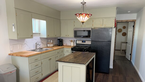 Una cocina compacta con armarios verde claro, encimera de madera, horno empotrado, refrigerador negro y una lámpara colgante.