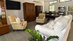 Una sala de estar con un sofá color beige, dos sillones, un aparador de madera, un televisor de pantalla plana y una zona de cocina con microondas y refrigerador.