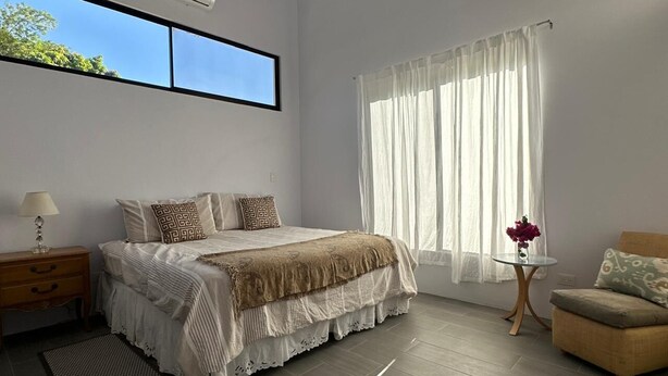 Un dormitorio con cama, mesita de noche, silla, una pequeña mesa con un jarrón y ventana con cortinas.