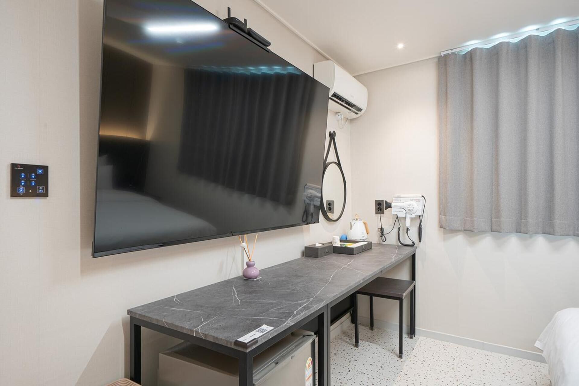 A modern hotel room with a large flat-screen TV mounted on the wall, a desk with a chair, a hairdryer, and a mirror.