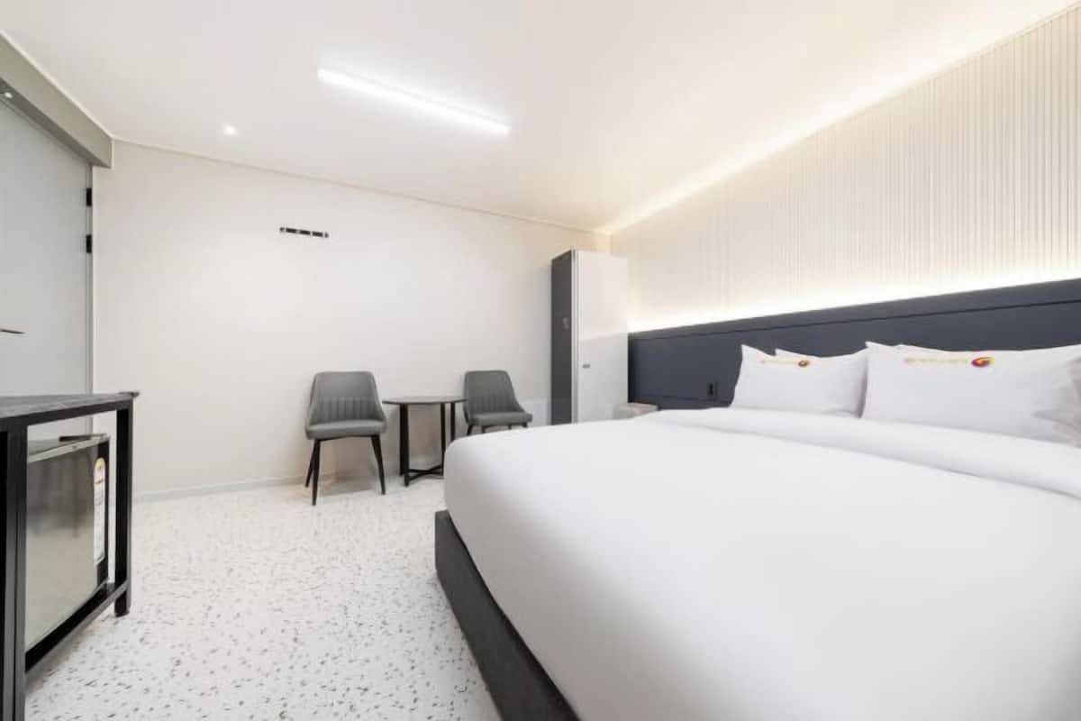 A modern hotel room with a large bed, a desk, two chairs, and a minibar.