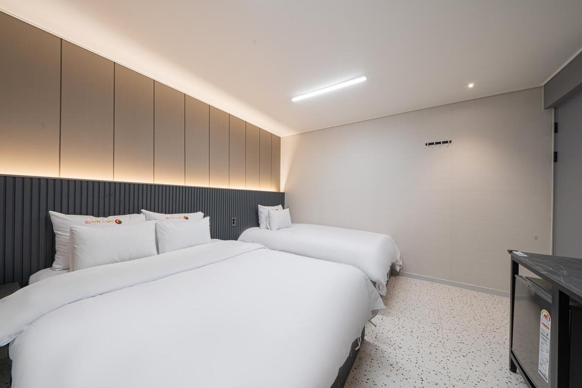 A modern hotel room with two beds, a dark headboard, a small desk, and a ceiling light.
