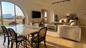 A modern living room with a glass dining table, black wicker chairs, a beige sofa, and a large window offering a view of a Ferris wheel and buildings. - Large 5br, Sea & JBR view, Easy Beach Access, Family Friendly! (Dubai)