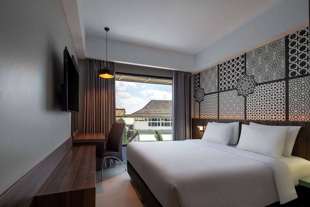 Ein modernes Hotelzimmer mit einem großen Bett, einem Schreibtisch mit Stuhl, einem an der Wand befestigten Fernseher und Blick auf ein Gebäude.