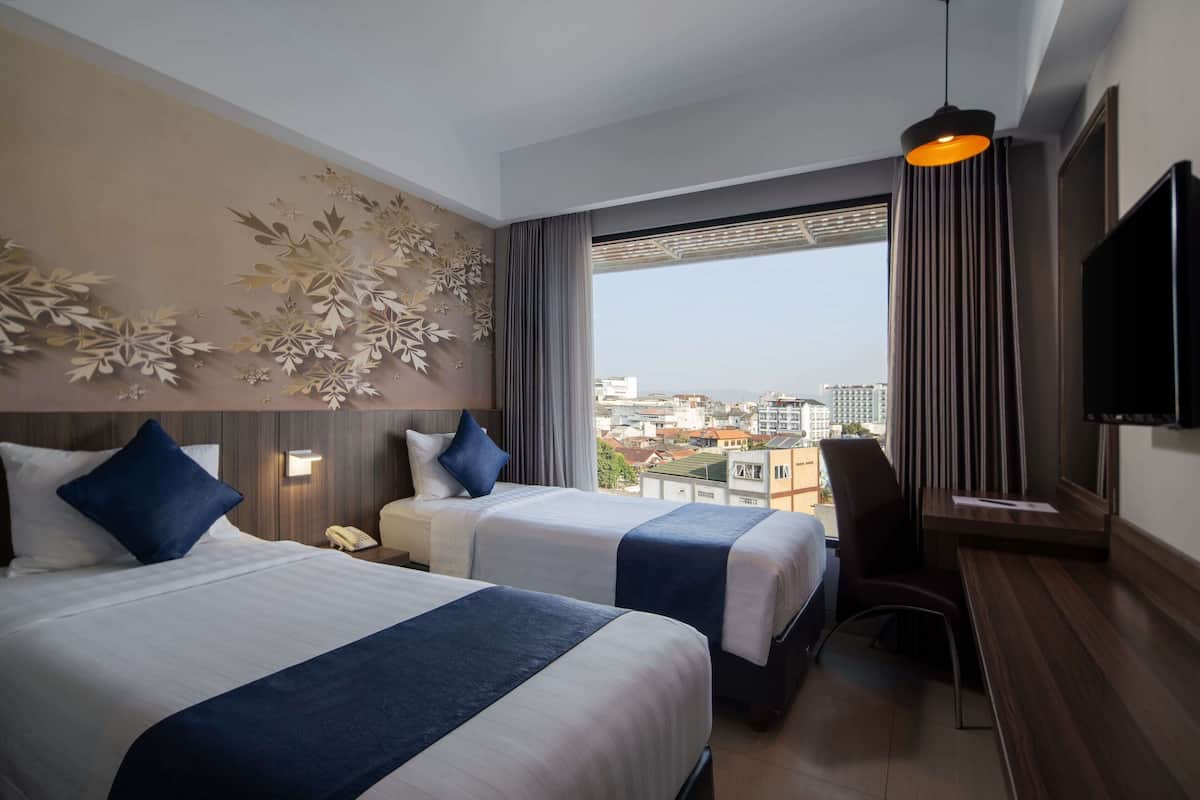 Ein Hotelzimmer mit zwei Betten, einem Schreibtisch, einem Fernseher und einem großen Fenster mit Blick auf die Stadt.