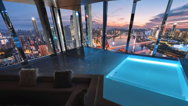 Ett modernt hotellrum med ett stort fönster som erbjuder utsikt över staden, ett glasbord och en soffa med fluffiga kuddar.