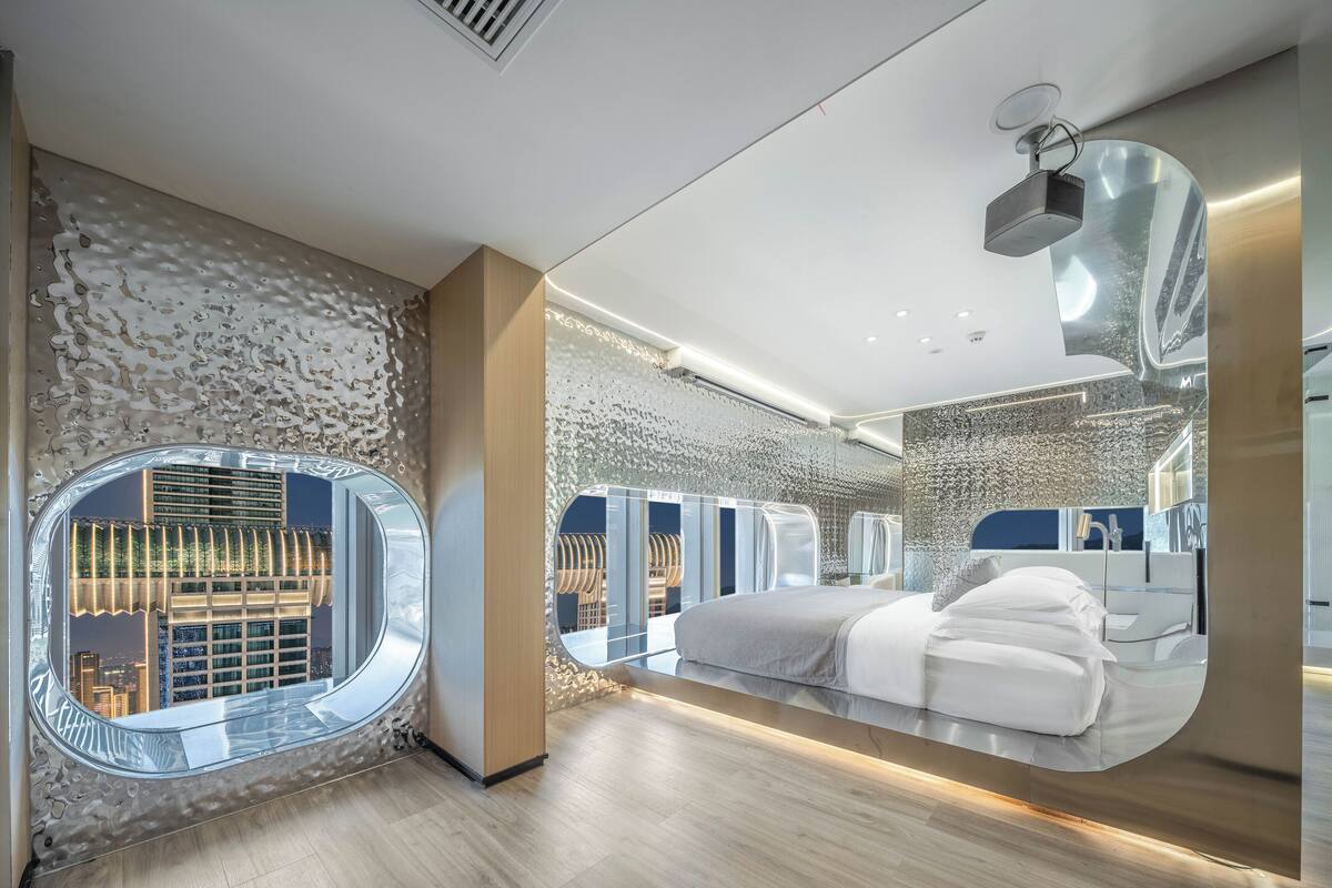 A modern hotel room with a large mirror, a bed, a bathroom, and a view of a cityscape through a circular window.