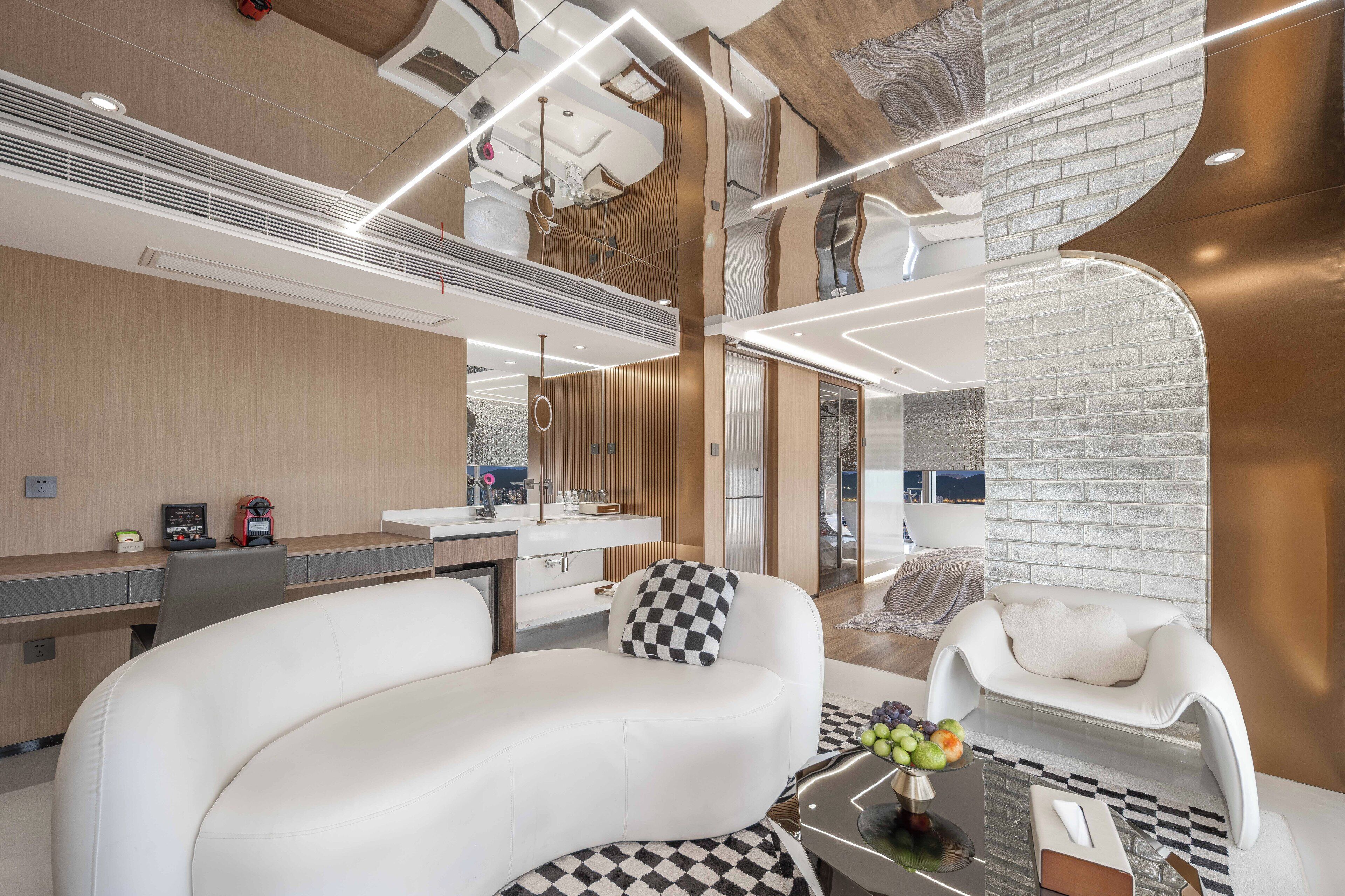 Una habitación de hotel moderna con un sofá blanco curvo, una mesita de centro redonda con fruta, un sillón blanco y una alfombra a cuadros.