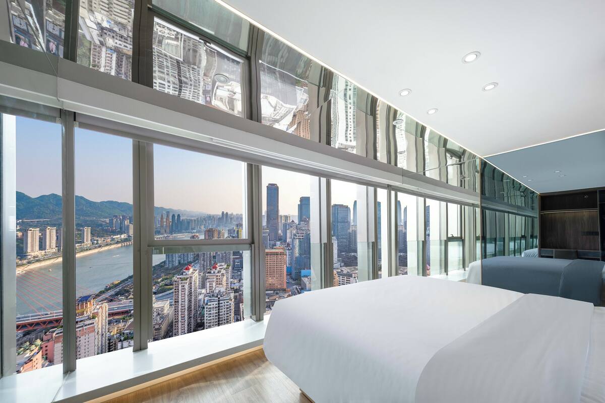 A modern hotel room with a large bed, a city view through floor-to-ceiling windows, and a minimalist design.