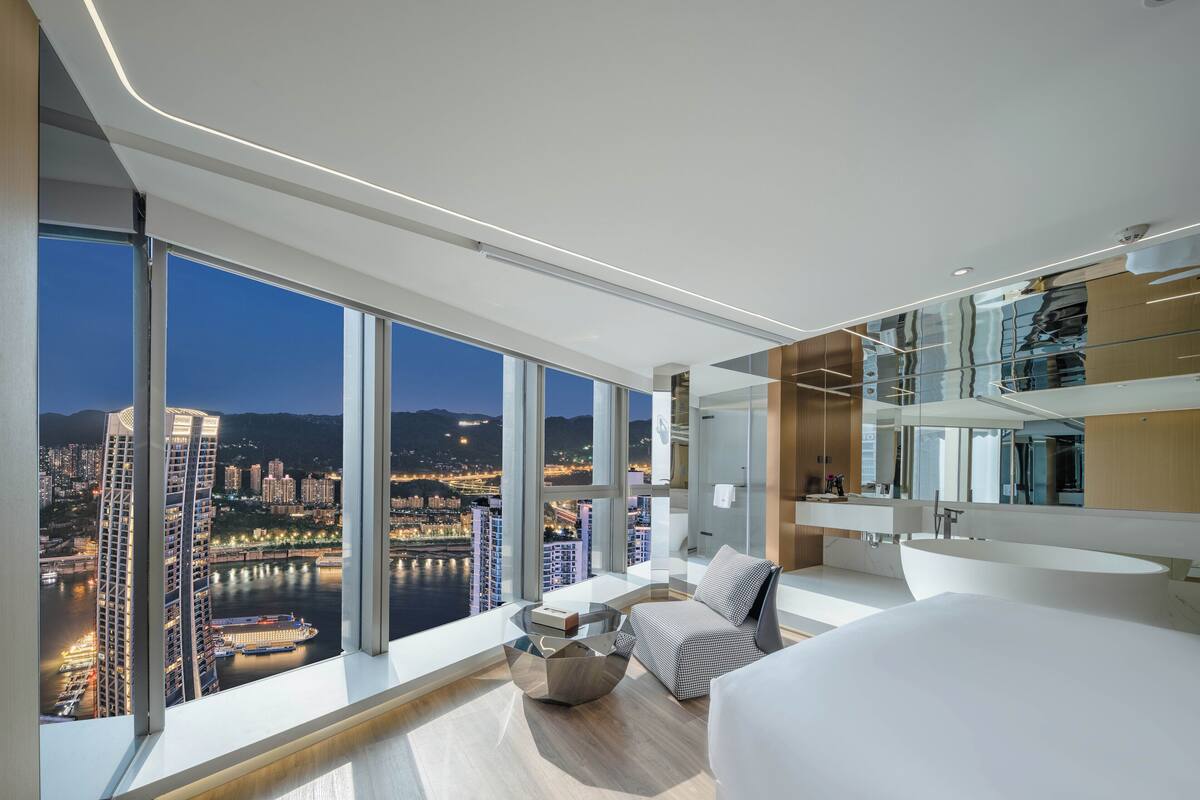 A modern hotel room with a large city view, a cozy seating area, and a minimalist design.