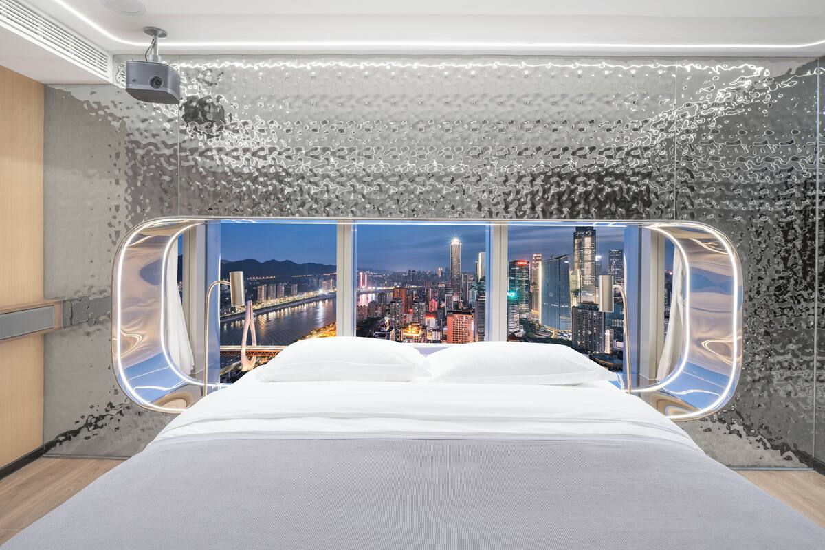 A modern hotel room with a large bed, a city view through a unique window design, and a ceiling-mounted projector.