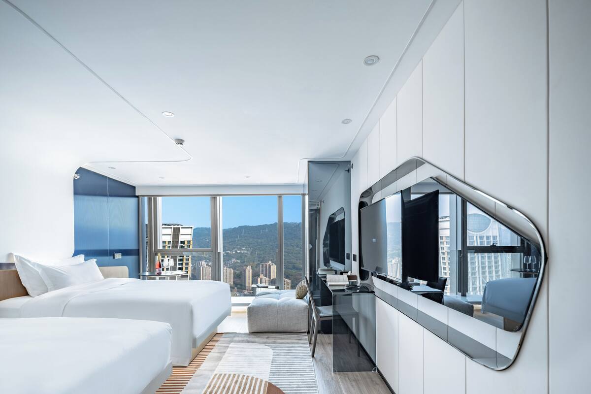 A modern hotel room with two beds, a large mirror, a desk, and a view of the city.