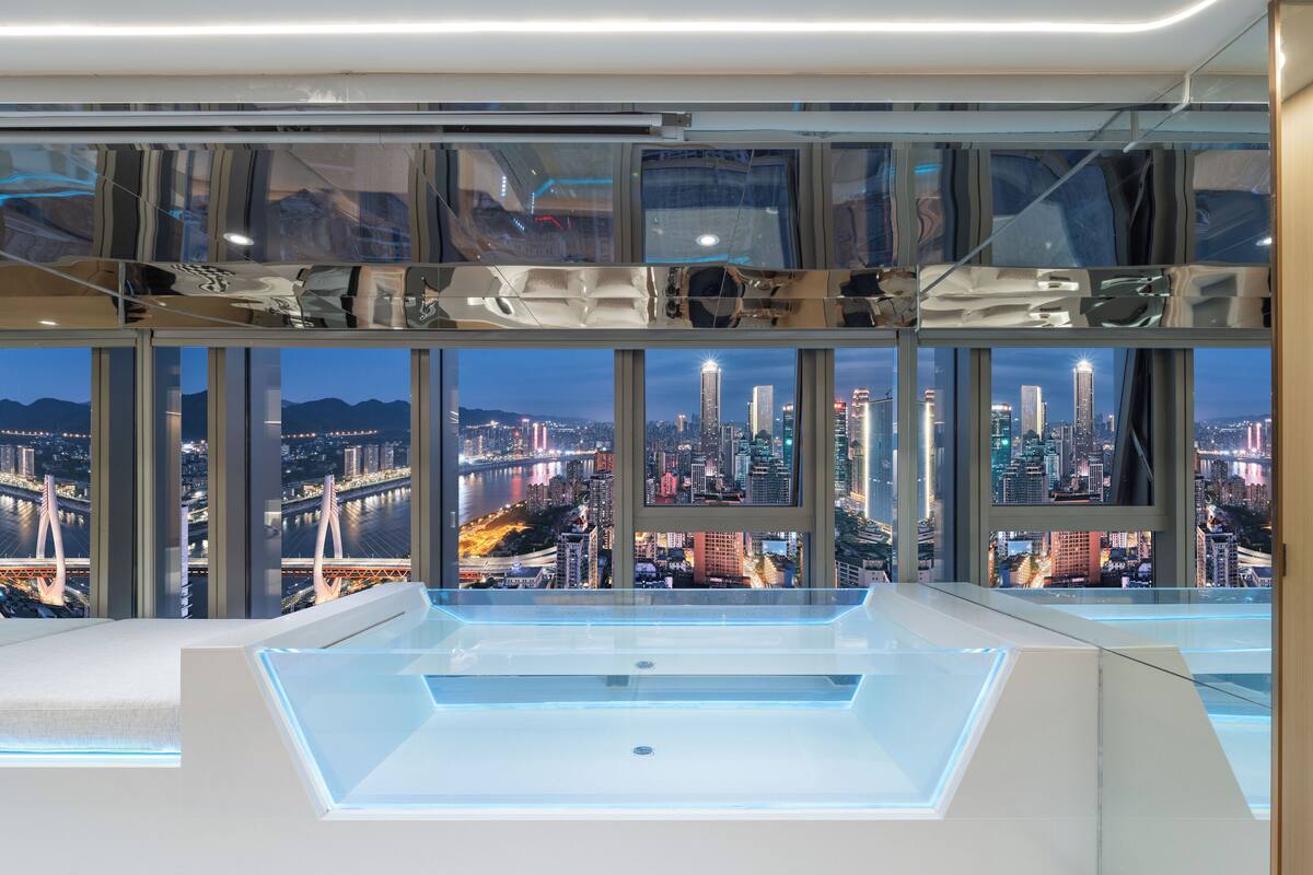 A modern hotel room with a large city view through floor-to-ceiling windows, a minimalist design, and a central water feature.
