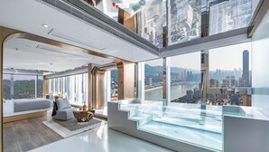 Ett modernt hotellrum med en glaspool som ser ut att sträcka sig oändligt, utsikt över staden och en mysig sittgrupp med soffa och soffbord.