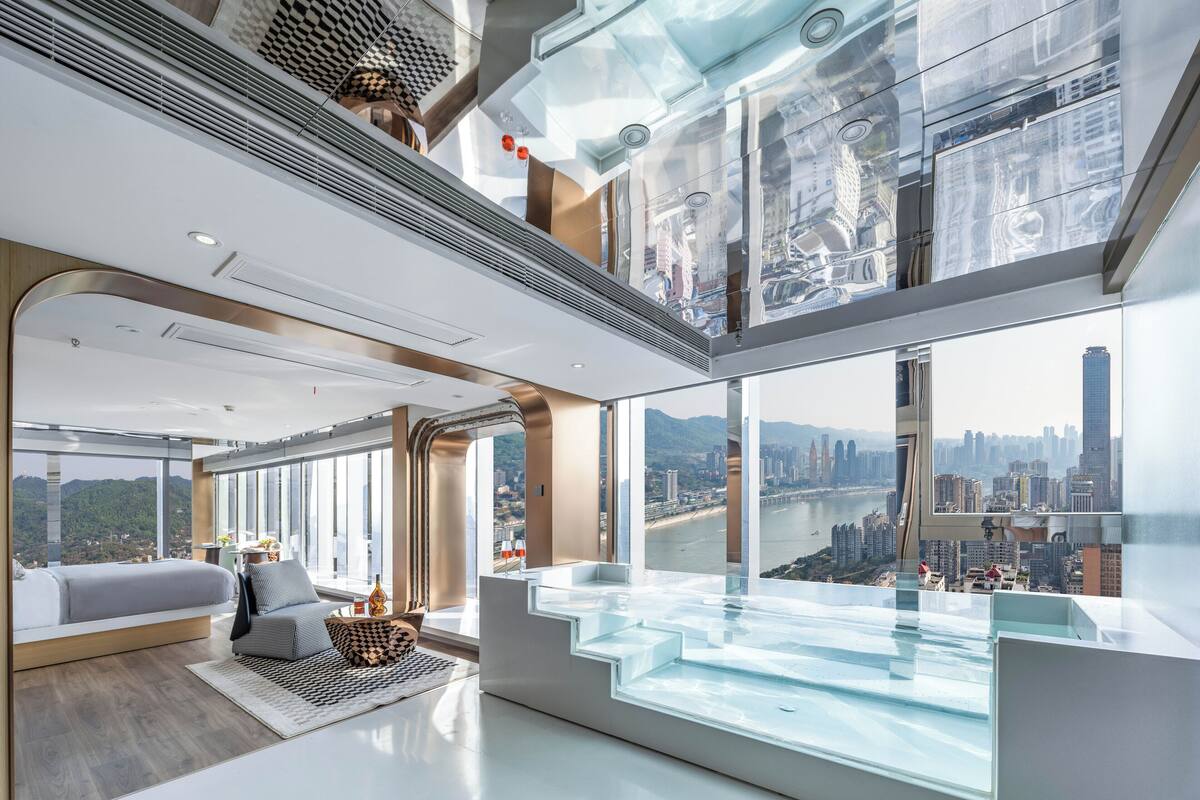 A modern hotel room with a glass infinity pool, a city view, and a cozy sitting area with a sofa and coffee table.