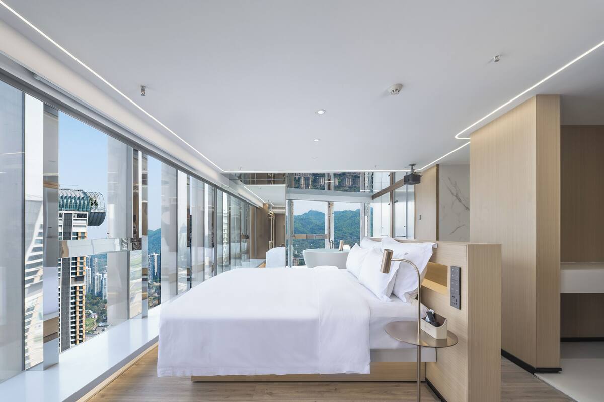 A modern bedroom with a large bed, a bedside table, and a view of a cityscape through floor-to-ceiling windows.