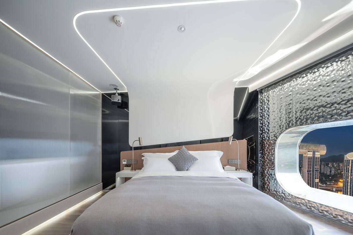 A modern hotel room with a large bed, a view of the city through a window, and a textured wall design.
