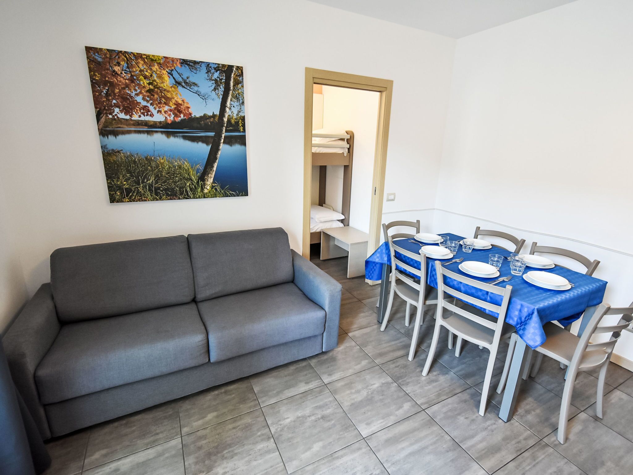 A living room with a grey sofa, a dining table with blue tablecloths, and a painting of a lake.